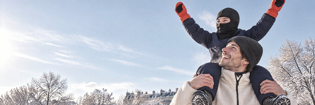 Enfant en cagoule sur les épaules de son père, bras levés, moufles orange