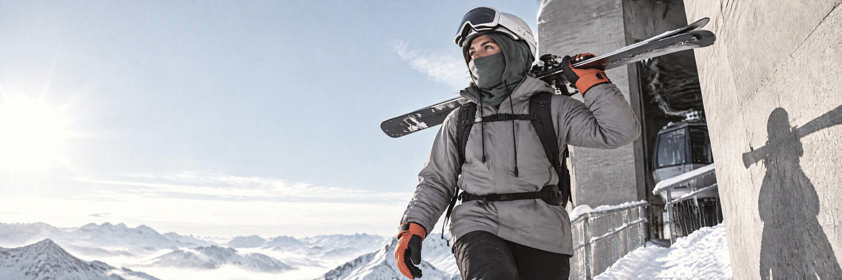 Skieuse skis sur l'épaule devant une gare de téléphérique, cagoule de ski et gants orange