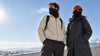 Un homme et une femme sur un toit face aux montagnes, cache-cou et cagoule noirs, bonnet orange